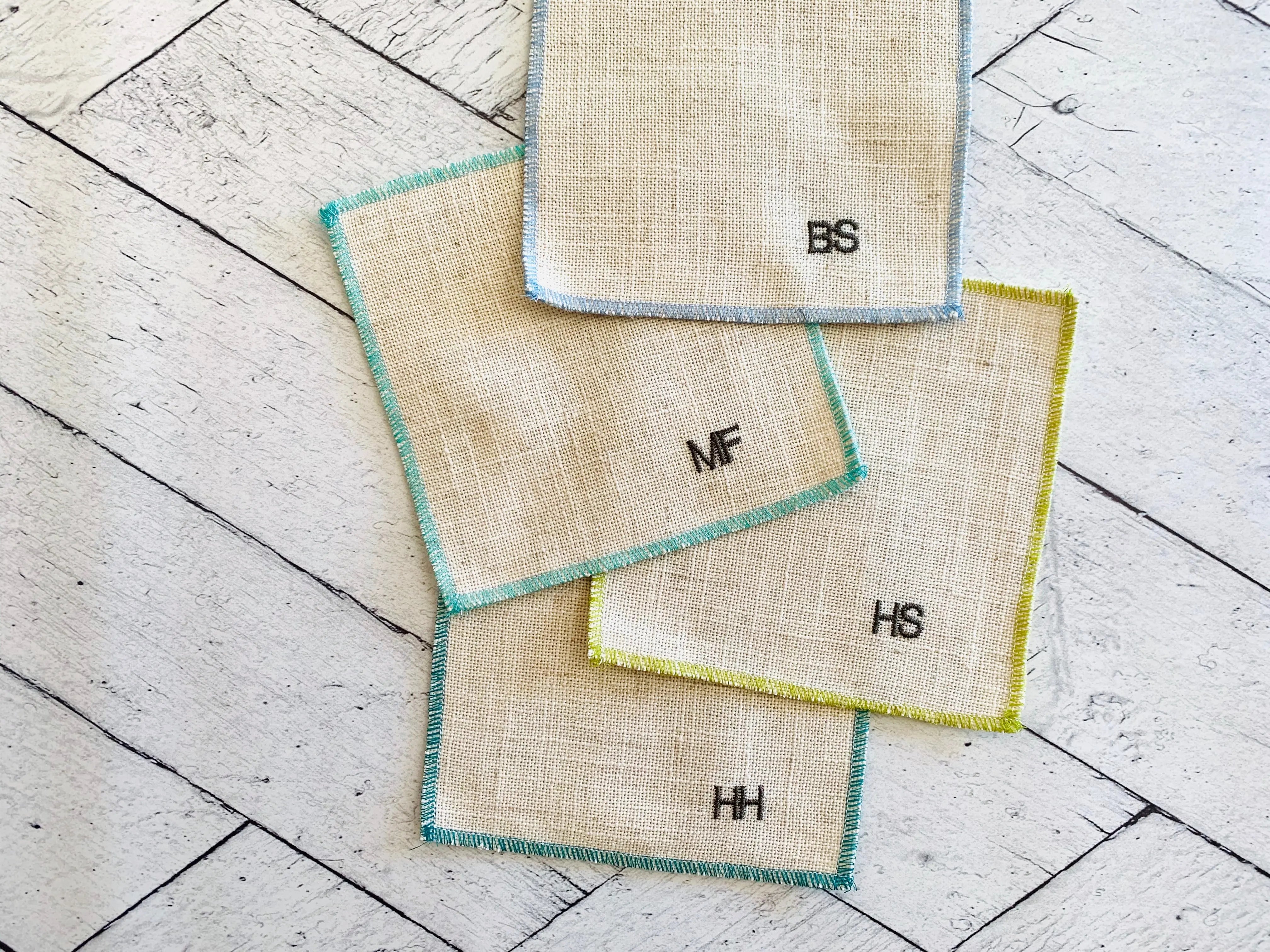 Linen cocktail coasters with monograms BS, MF, HS, and HH on a wooden background.