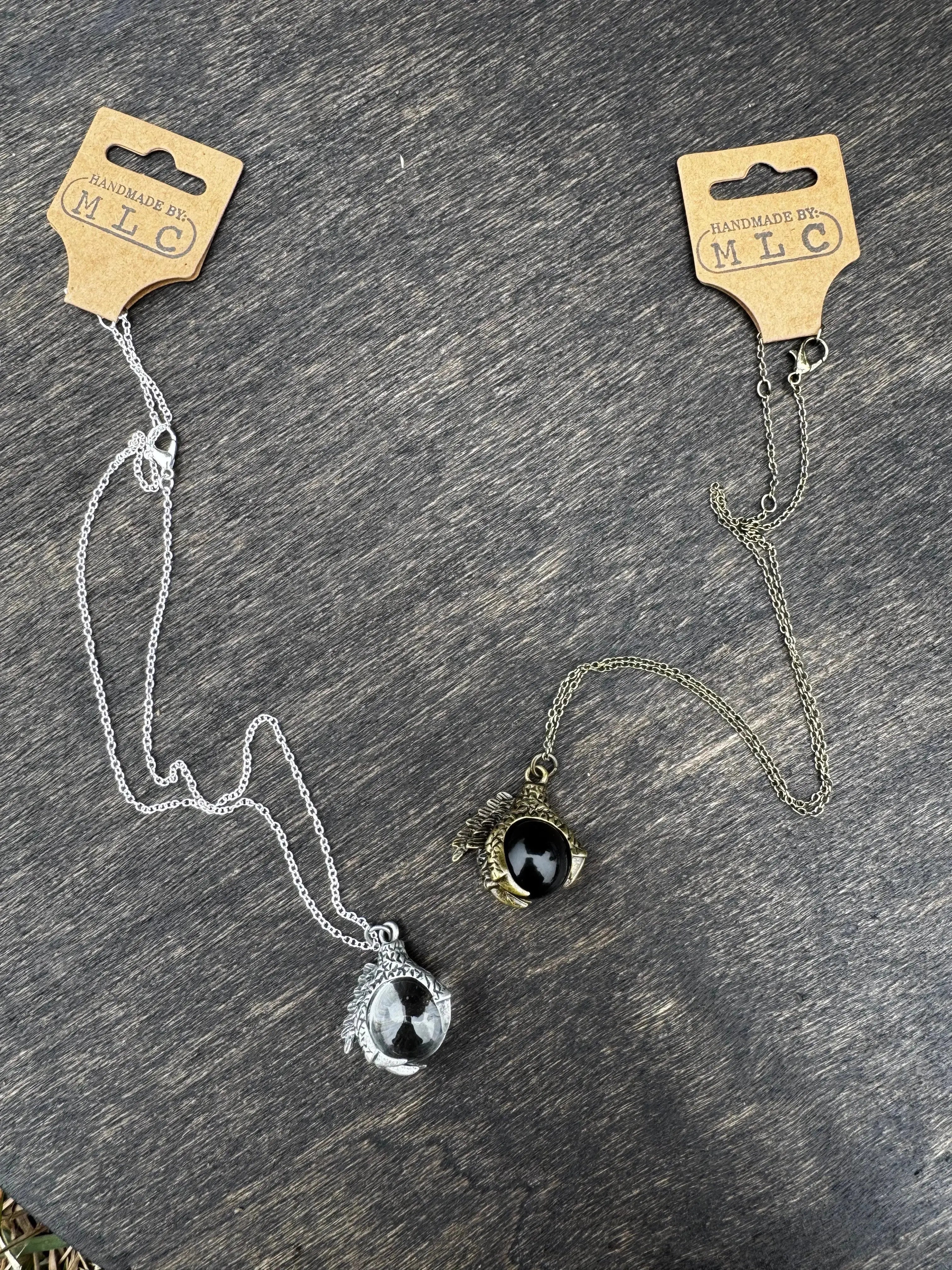 Two dragon claw necklaces on a wood surface. One silver with a clear orb, the other gold with a black orb.