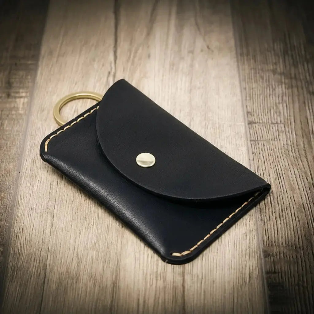 Black leather keychain wallet with brass ring and snap closure on a wooden surface.