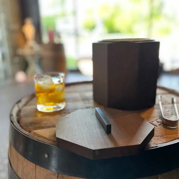 Blackened cherry wood ice bucket with lid and whiskey glass on a wooden barrel. 