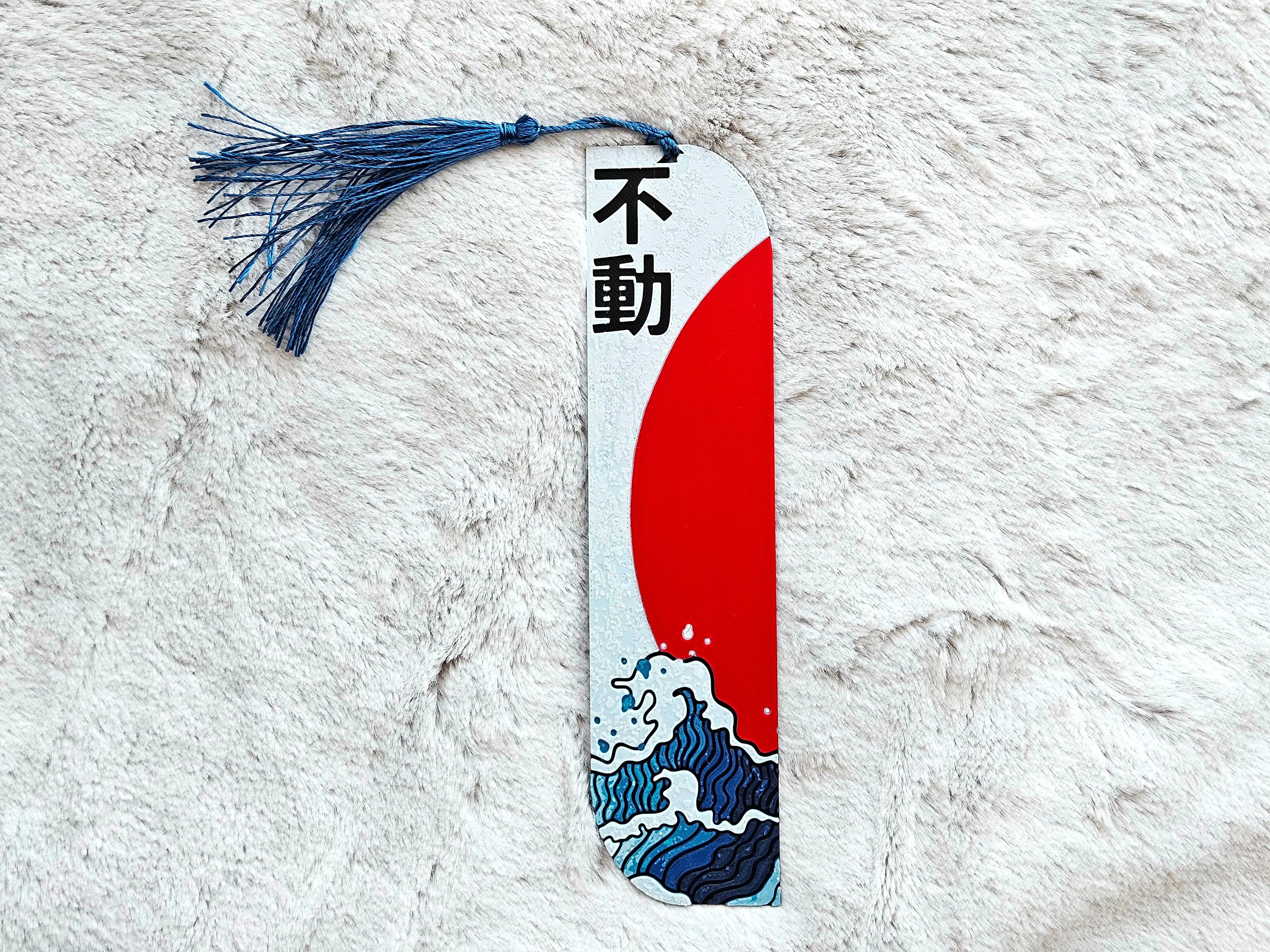Japanese bookmark with kanji, red sun, blue waves and tassel on a white textured surface.