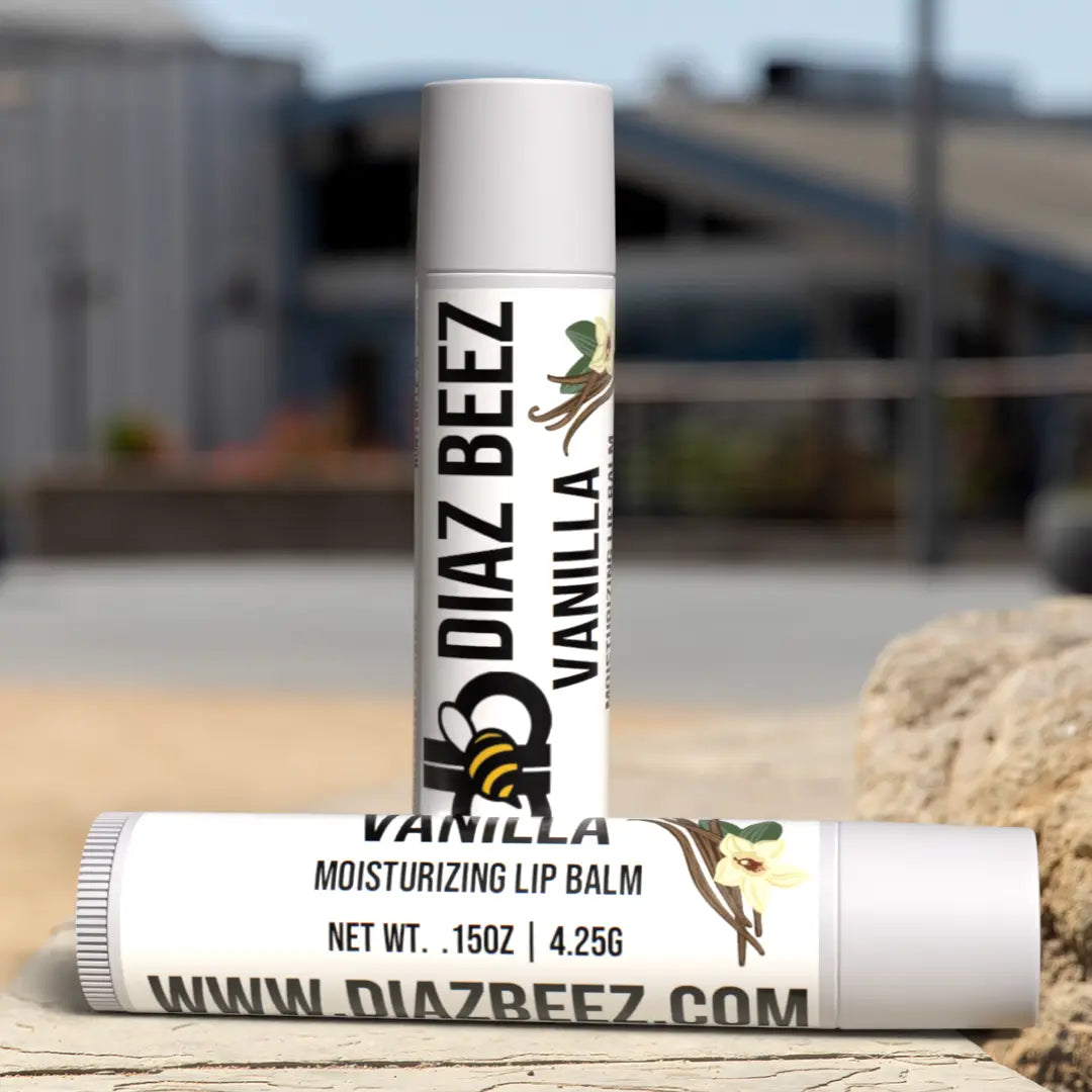 Two Diaz Beez vanilla lip balm tubes, one standing and one laying down on a textured surface, with outdoor background.