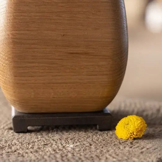 White oak wooden vase with a bronze base and a small yellow flower on a brown surface.