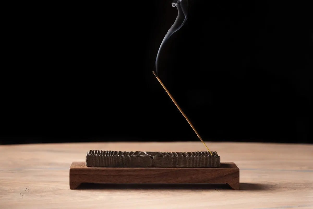 Incense stick burning in a wooden holder with bronze casting detail and smoke rising against a black background.