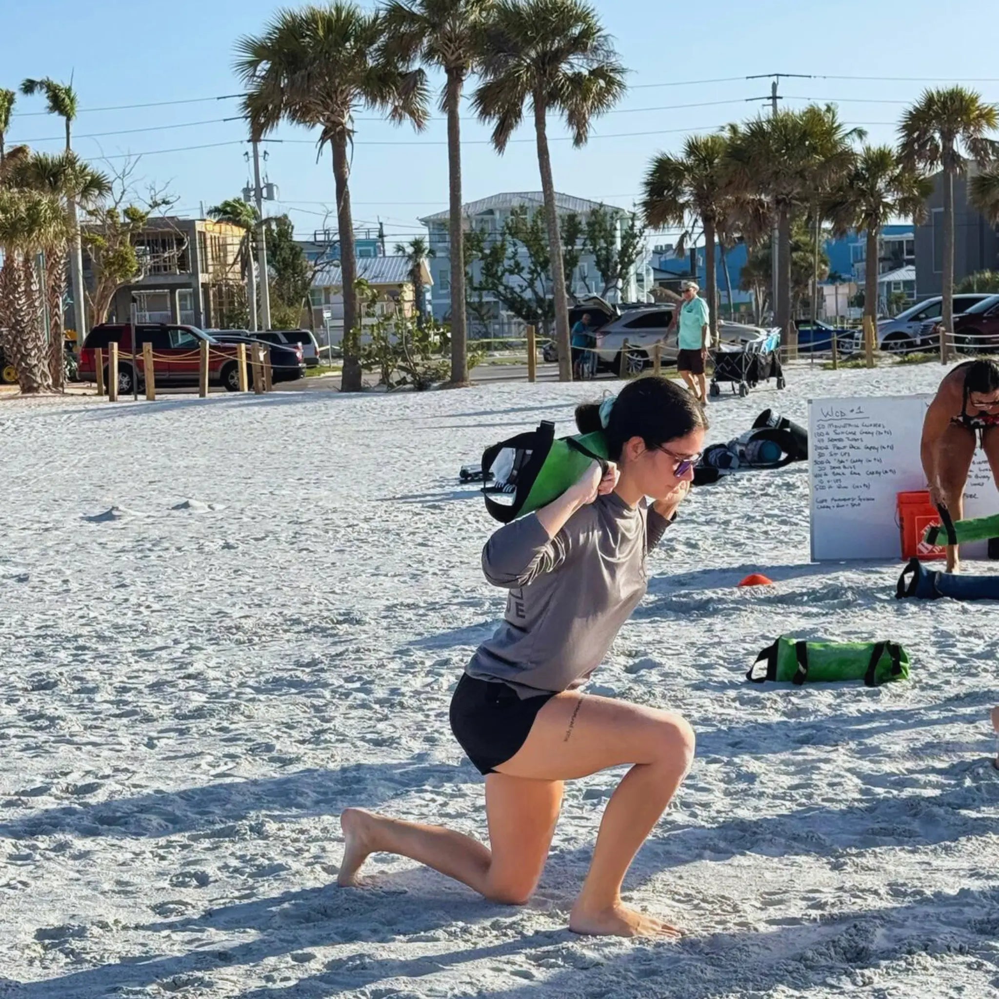 People exercising with green sandbags on a sandy beach with palm trees.